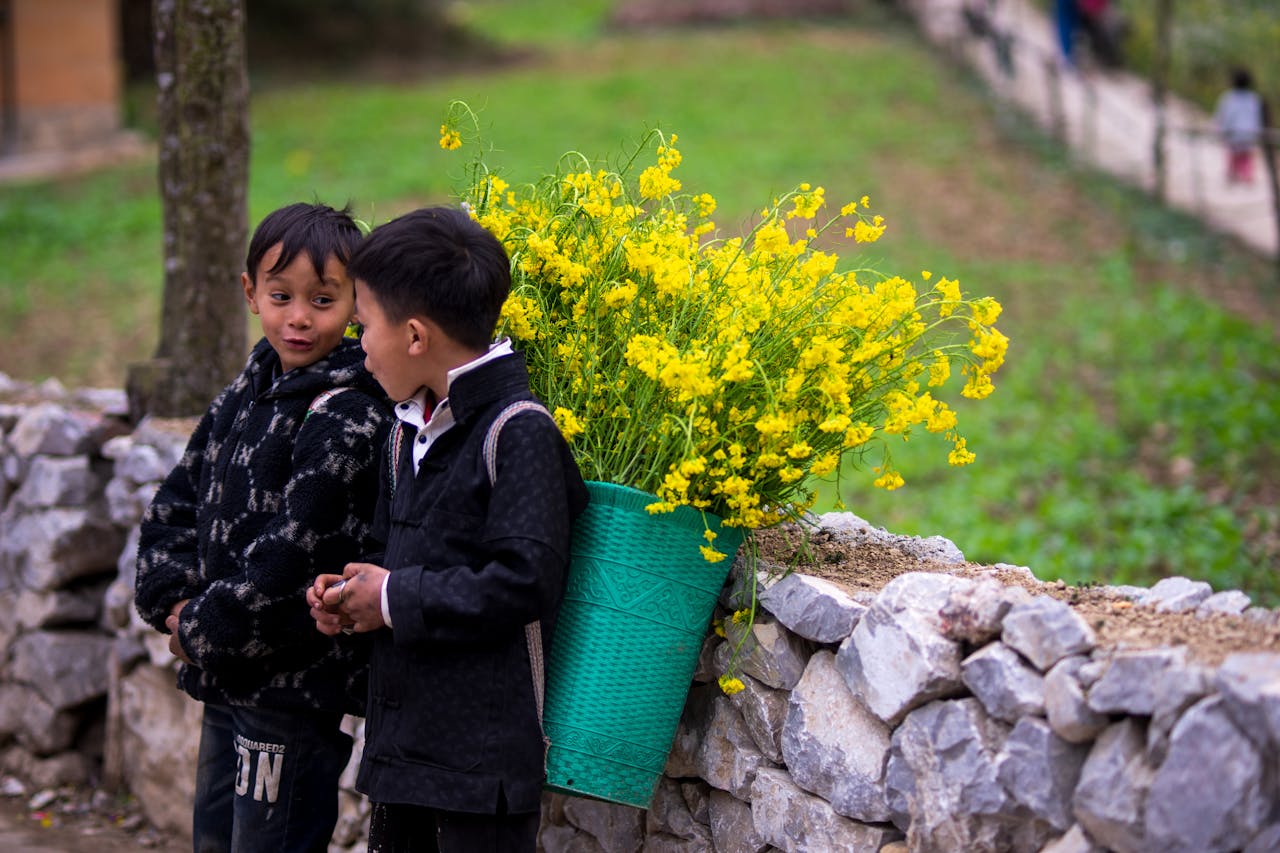 Two children in Ha Giang, Vietnam, with a basket of yellow wildflowers by a stone wall.
